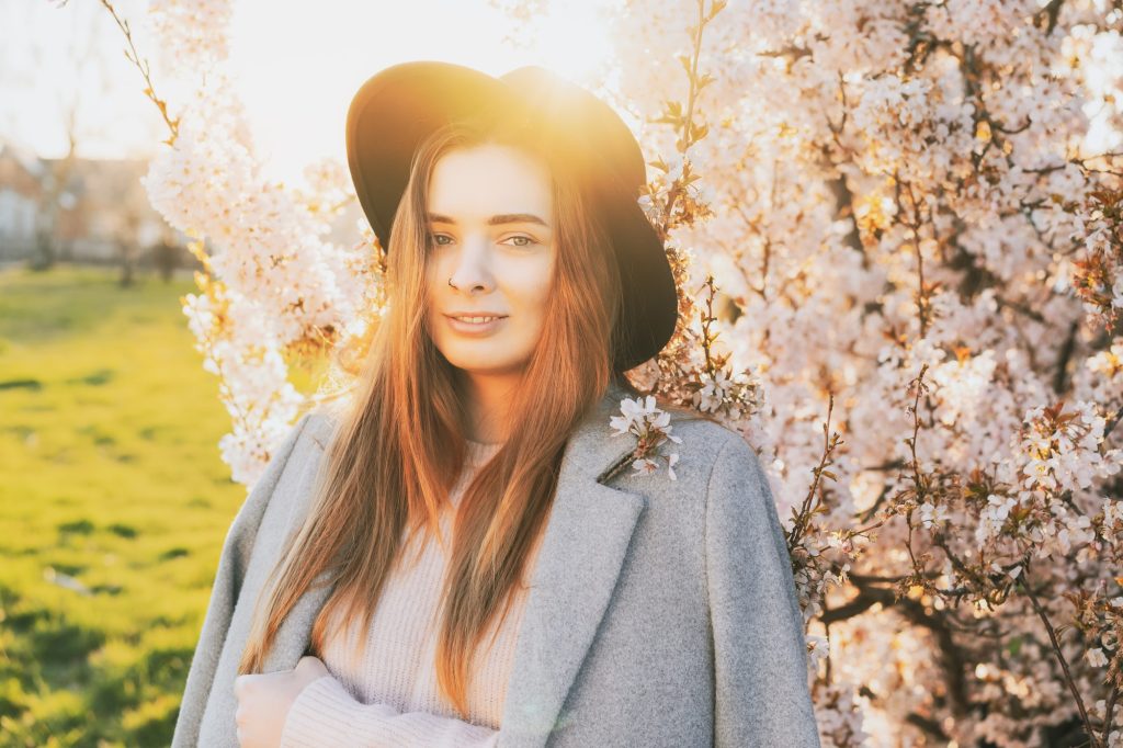 Portrait of a young woman in hat and coat near blooming tree with sunset light and sunbeams. Gen Z